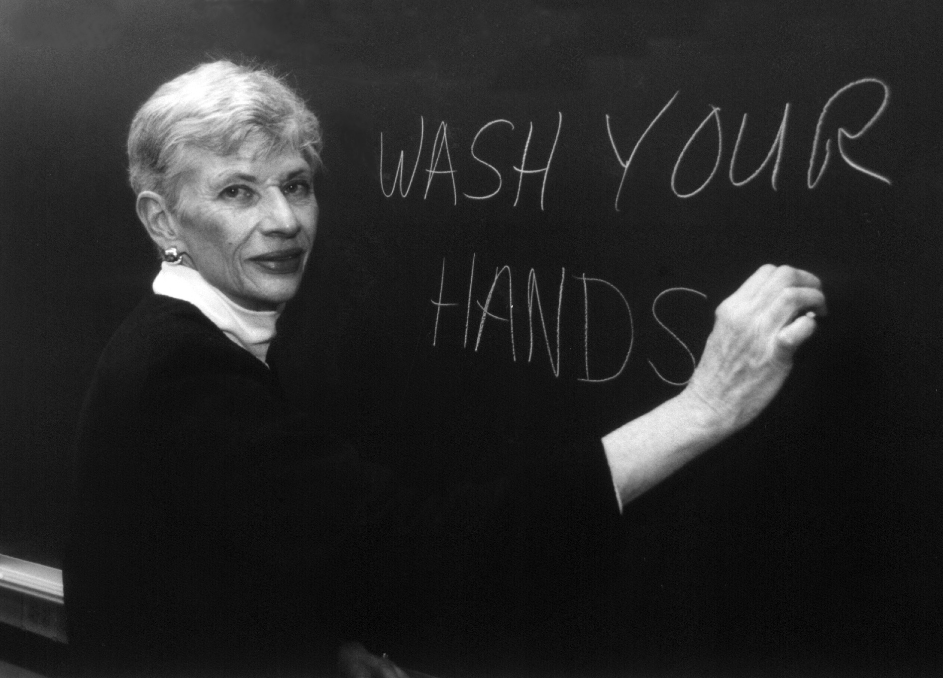 Microbiologist Dixie Whitt writes on a chalkboard, "Wash your hands."