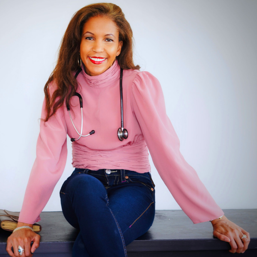 Nicole Williams smiles while sitting atop a desk with a stethoscope draped around her neck.