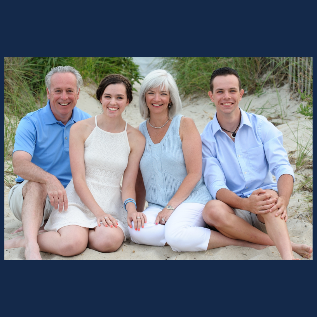 Illinois alumnus Michael Recny and his family (daughter, Brenna; wife, Cate; son, Donovan) sit together on a beach in various shades of light blue and white.