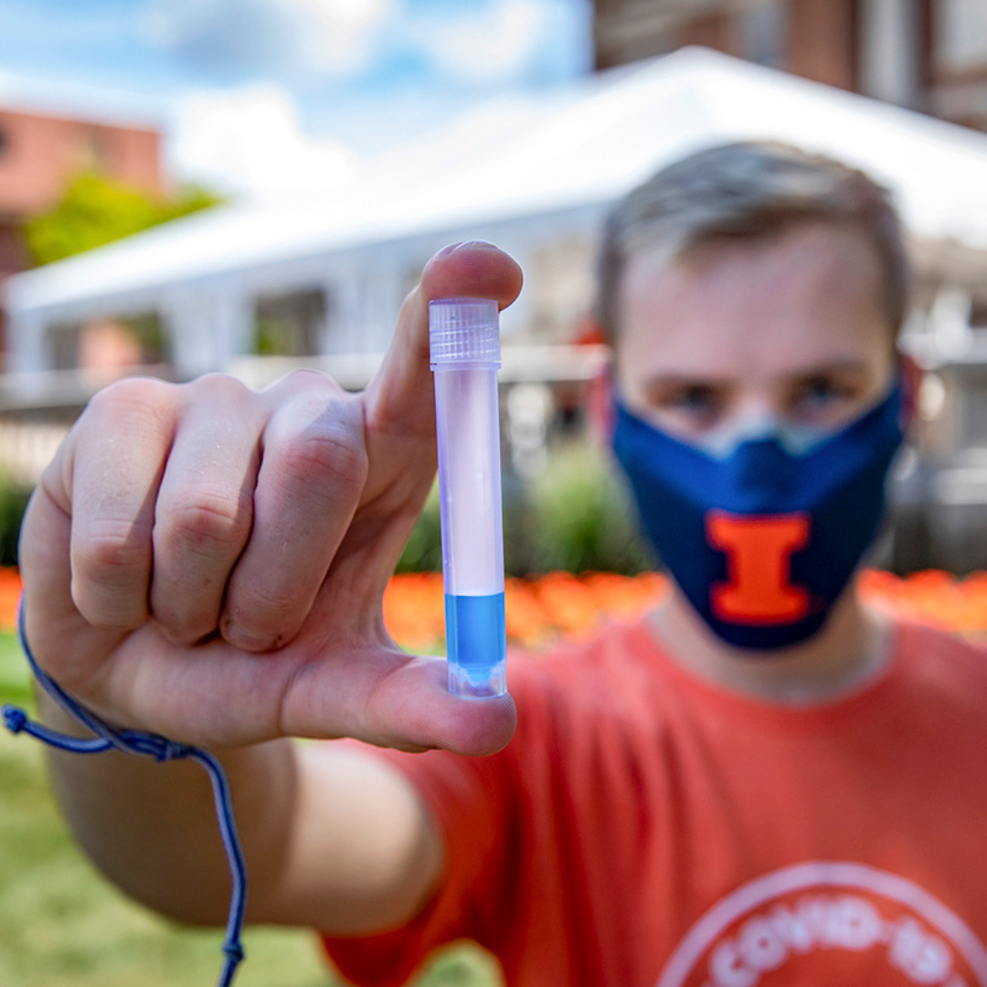 A student holds up a vial with saliva. The vial is sharply in focus, while the student, who is wearing a U of I face mask, is blurred out of focus. 