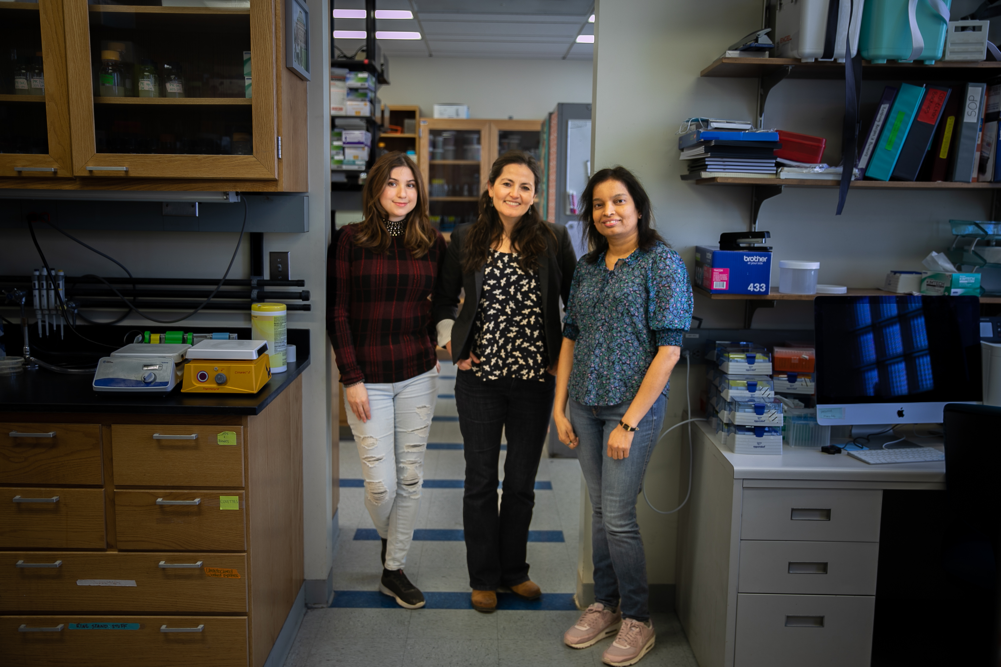 Three scientists in the Mera Lab, Stephanie Puentes-Rodriguez, Paola Mera, and Inoka Menikpurage, stand together smiling for the camera. They are in the doorway leading from one lab space to additional lab space. They are all smiling.