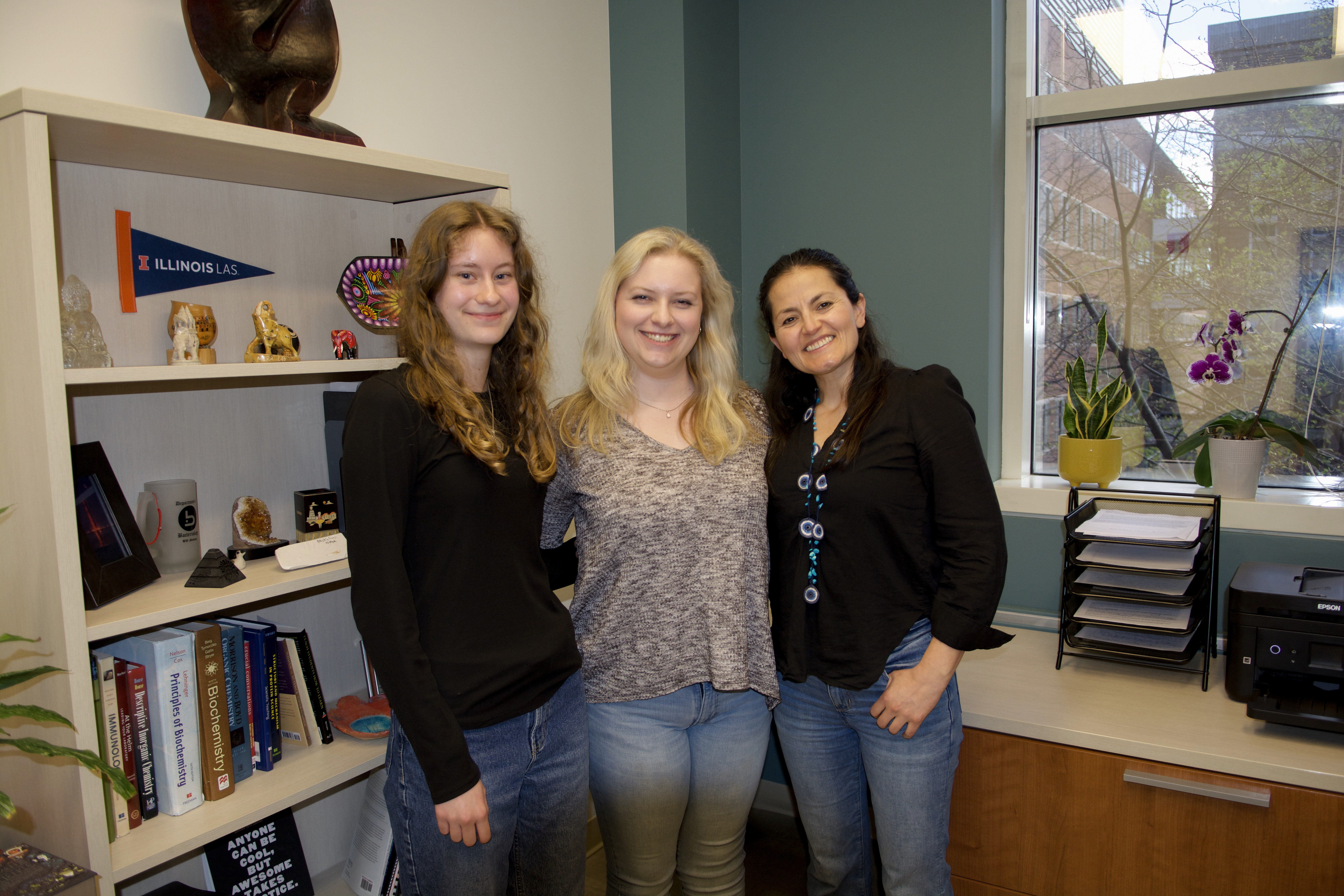 Three women smile while standing in front of a bookcase.