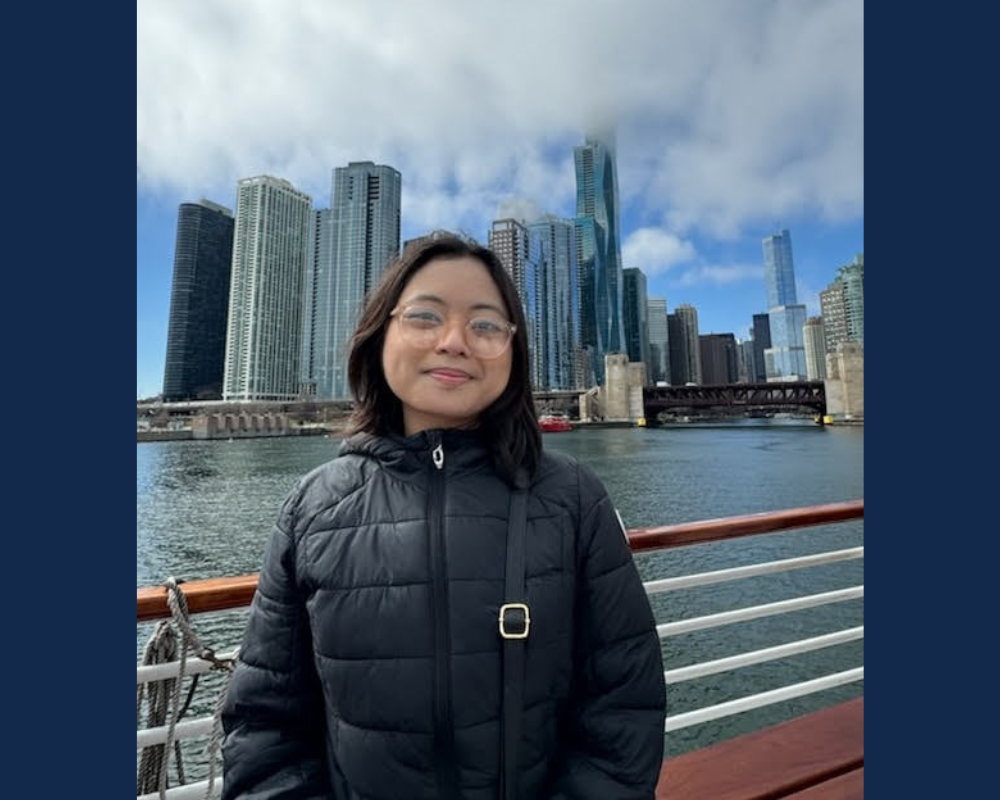 A woman in a black coat and glasses stands in front of a building skyline.