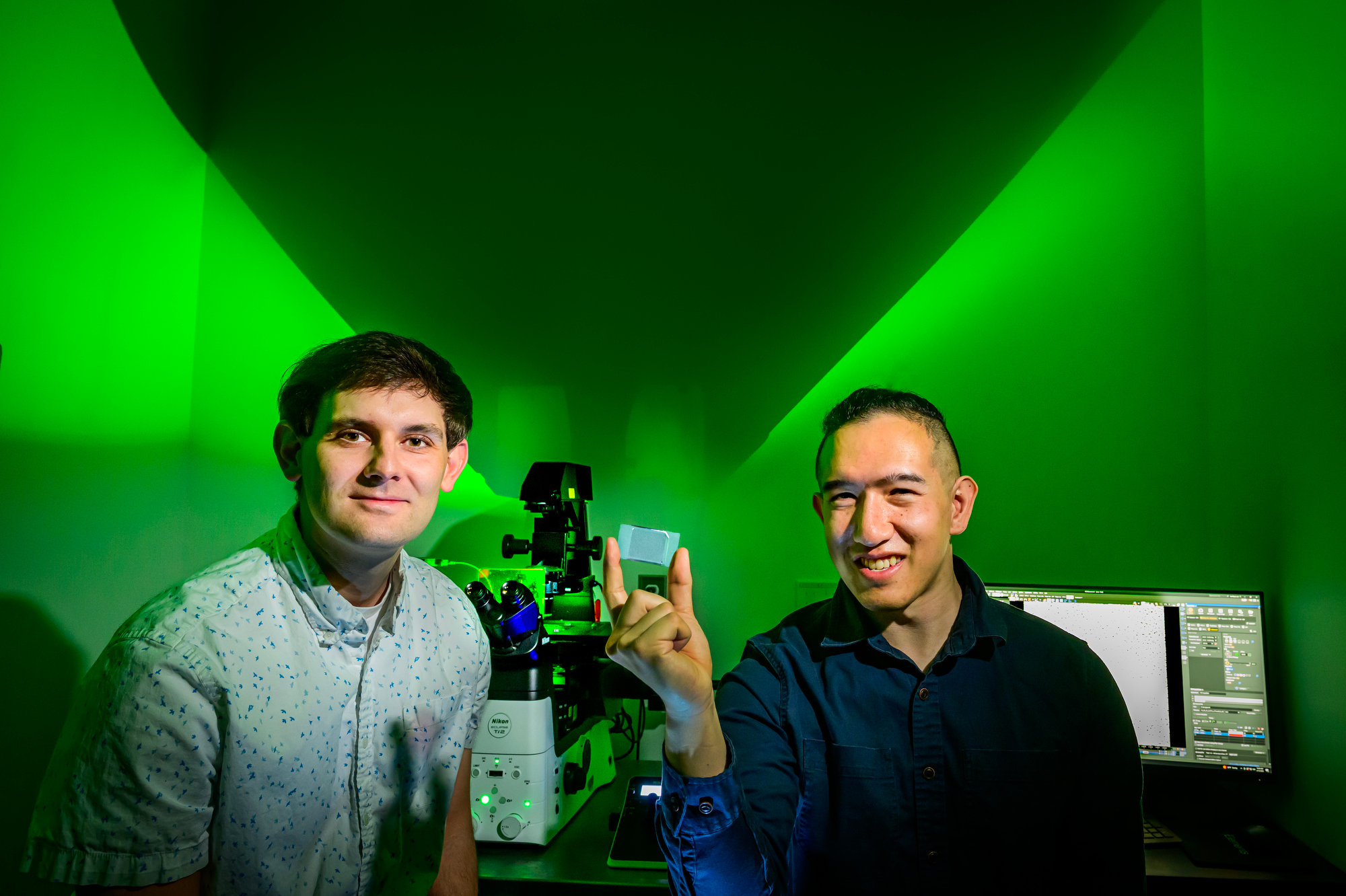 Researchers hold a microfluidic device while sitting in front of a microscope