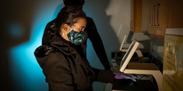Teaching assistant stands in lab looking at monitor. She is masked and her shadow has a blue tone.