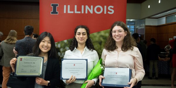 Three MCB students stand together for a group photo with MCB Honors certificates with a University of Illinois banner in the background.