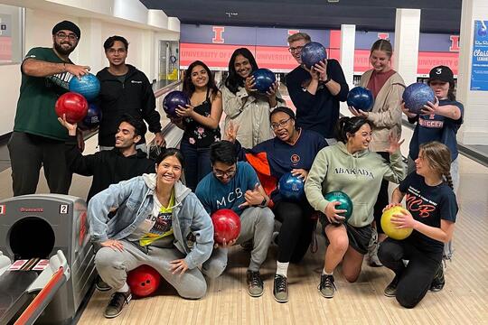 Roughly a dozen students (MCB Leaders) pose for the camera at a bowling alley, all holding bowling balls. Some are smiling at the camera, others are making silly expressions at one another. 