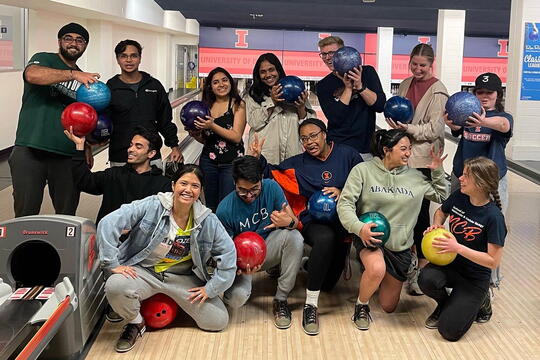Roughly a dozen students (MCB Leaders) pose for the camera at a bowling alley, all holding bowling balls. Some are smiling at the camera, others are making silly expressions at one another.