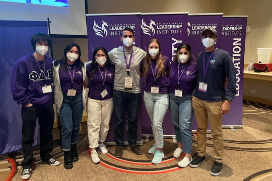 Seven students stand in a row with arms around each other, all wearing masks and name tags. Banners for "Phi Delta Epsilon Leadership Institute" are behind them.