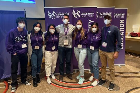 Seven students stand in a row with arms around each other, all wearing masks and name tags. Banners for "Phi Delta Epsilon Leadership Institute" are behind them. 