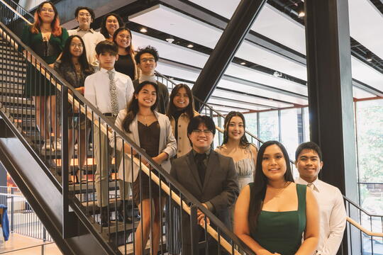 Roughly a dozen students stand in two lines on a staircase, all facing the camera and smiling. 