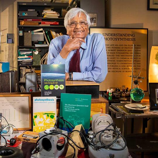 Professor Govindjee smiles for the camera in his office, where books and equipment are on display on a table in front of him. Behind him, a plaque titled "Understanding Photosynthesis" is propped up.