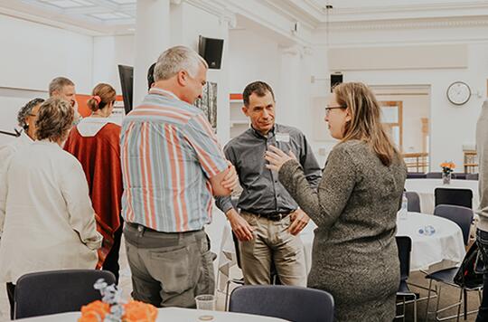 Three faculty members stand talking to one another in between tables set up at a faculty event. 
