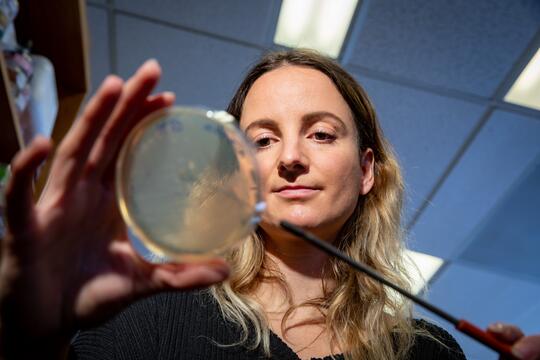 Stefanie Eben holds up a Petri dish. The dish is in soft focus, with Stefanie in sharp focus behind it. 