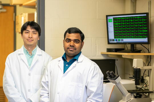 Two researchers in white lab coats stand in front of a computer monitor.