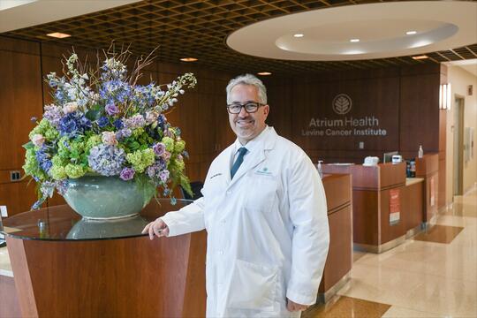 A smiling man in a white lab coat stands in front of a flower arrangement.