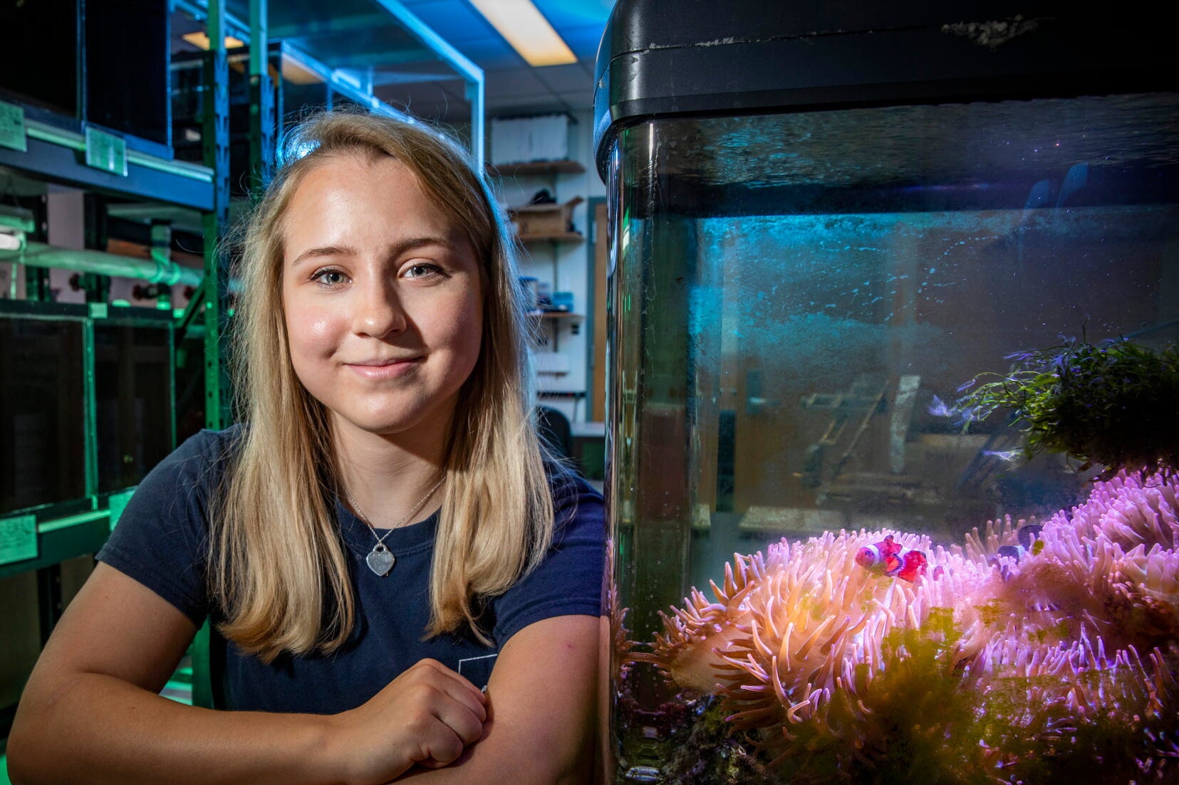 Student researcher poses in lab near aquarium of coral.