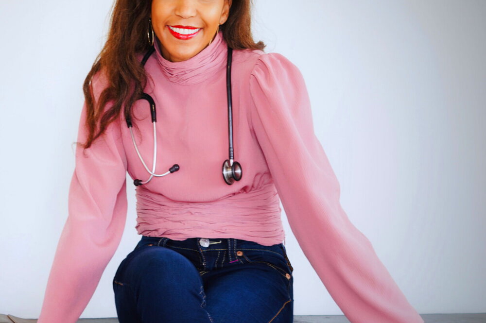 Nicole Williams smiles while sitting atop a desk with a stethoscope draped around her neck.