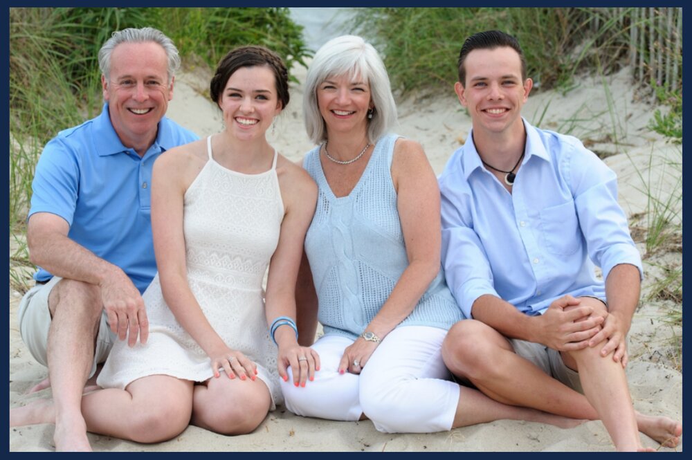 Illinois alumnus Michael Recny and his family (daughter, Brenna; wife, Cate; son, Donovan) sit together on a beach in various shades of light blue and white.