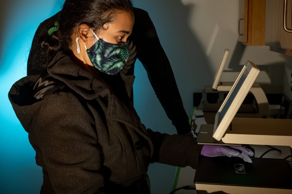 A graduate teaching assistant works in the lab with students looking over shoulder. She is silhouetted with a blue shadow. 