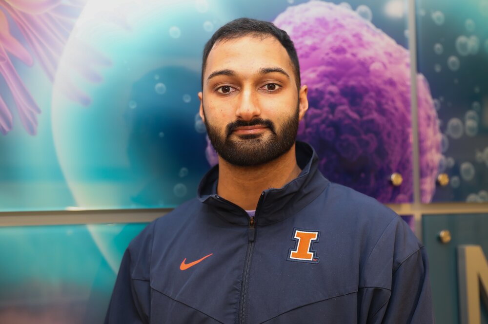 Headshot of Jay Sonalkar in front of a wall with illustrations of cells. He is wearing a U of I zip-up jacket.