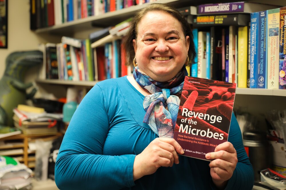 Microbiology professor Brenda Wilson stands in her office holding a copy of the book, Revenge of the Microbes