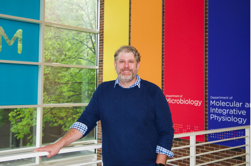 A man in a blue sweater smiles in front of a brightly-colored background