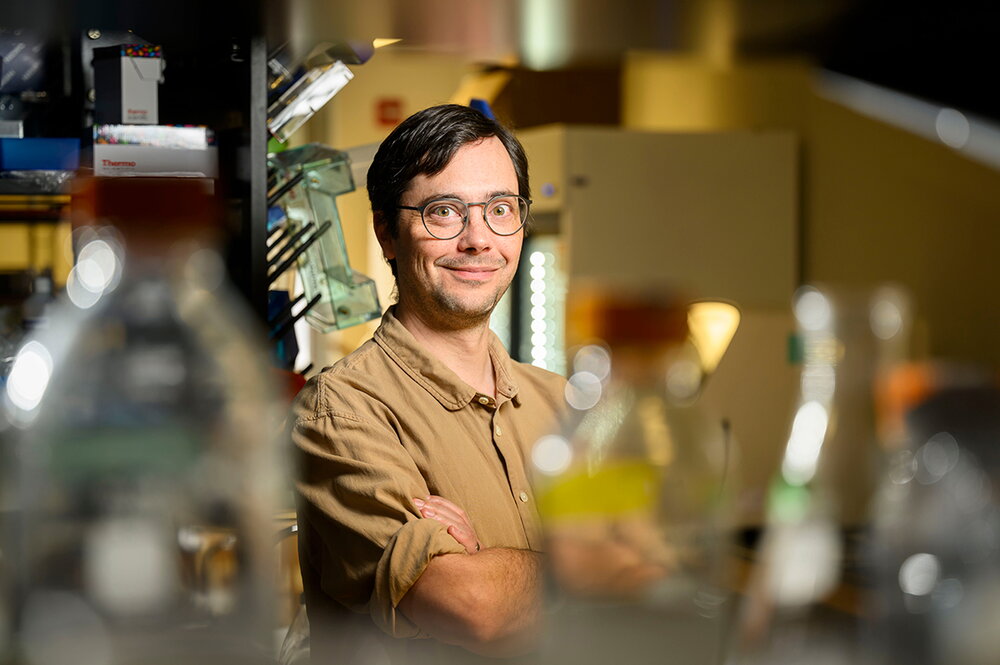 A man in a tan shirt and glasses stands in front of lab beakers.