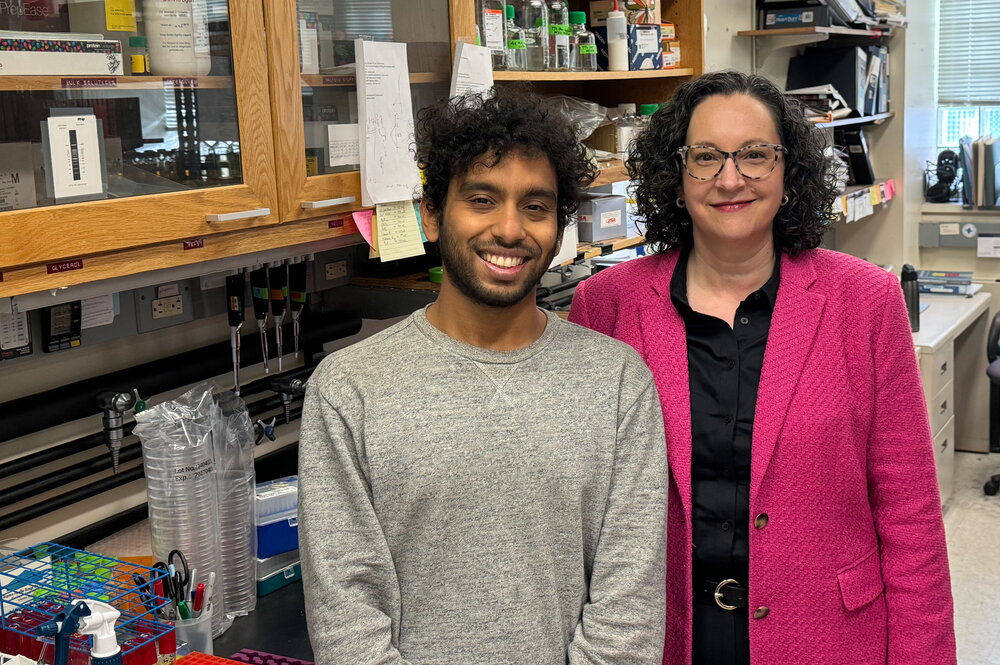 A woman in a pink blazer and a man in a grey sweater stand in a laboratory.