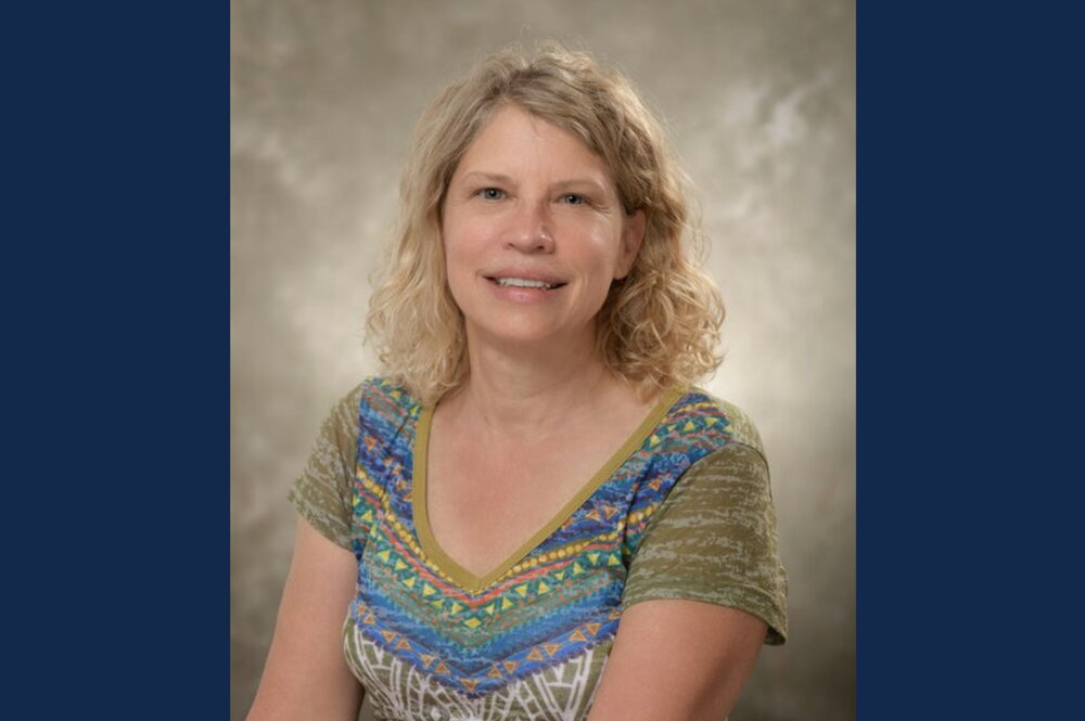 blond woman in green and blue shirt sits in front of beige backdrop