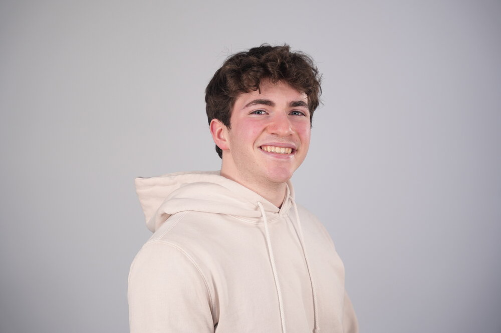 Man in cream colored sweatshirt poses for headshot in front of gray background