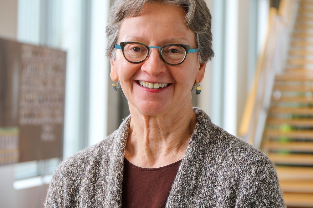 Woman wearing glasses, brown shirt and sweater poses in front of a stairway