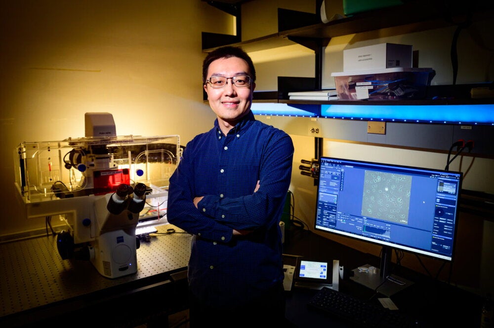 Wei Qin in blue shirt stands in front of computer screen and microscope