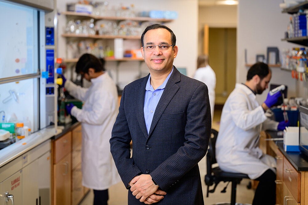 Wearing a blue blazer, Nash Kalsotra stands in a biochemistry lab between two benches while two graduate students behind him
