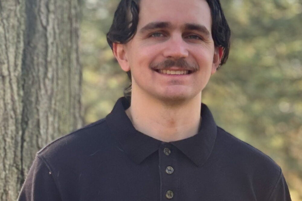 Man with brown hair and mustache stands outside wearing dark colored sweater