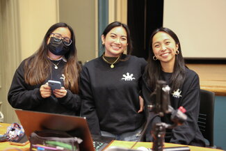 Three students stand together smiling in matching black t-shirts at the FACT conference. They are indoors.