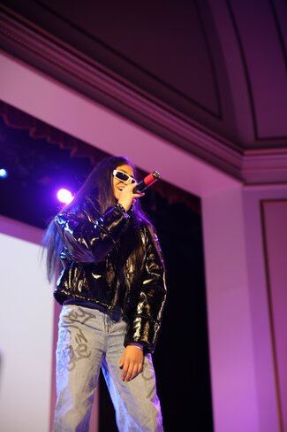 Ashley Mehta performs at the FACT Variety Show on stage. She is wearing a leather jacket and sunglasses indoors.