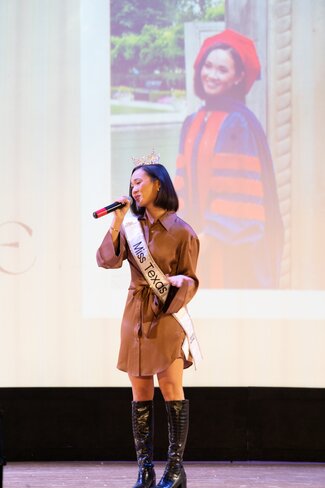 Keynote speaker Averie Bishop speaks on stage. She is wearing her Miss Texas sash and a picture of her in graduation regalia is up on a screen behind her. 