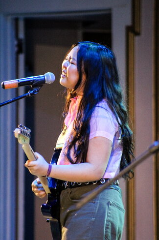 Ella Jay Basco performs at the FACT Variety Show. She is standing at a microphone singing and playing a guitar. 