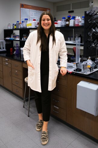 Madeleine Meehan stands in a lab by the bench, wearing a lab coat.