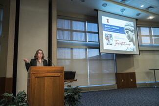 Rachel Whitaker speaks at the podium. A screen beside her says Investiture, with a picture of Whitaker outside IGB and a cutout picture of the Alma statue.