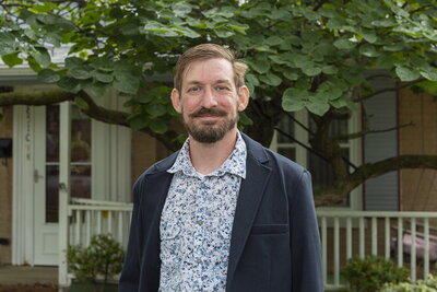 Man with beard and wearing blue blazer stands in front of porch