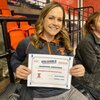 Jennifer Joesting holds up award certificate that says "Most Valuable Professor" while sitting in stands at UIUC women's gymnastics meet. 