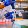 Gloved hands working with a pipette. Person's face can't be seen. They are in a lab coat and the background is a lab. 