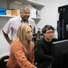 Drs. Kristen Flatt and Yiwu Zheng sit at a computer monitor. They are looking at the monitor, with Dr. Satish Nair standing behind them looking at the screen as well.