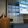 Whitaker speaks at the podium while a screen titled "Investiture of Rachel Whitaker" shows a picture of her and a cutout photo of Alma. 