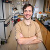 A brown-haired man in a tan shirt standing in a laboratory, posing with his arms crossed.