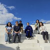 Six students sit on a white staircase in front of a blue sky.