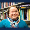 A smiling woman standing in front of a bookshelf wearing a blue shirt and scarf