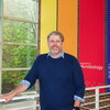 A man in a blue sweater smiles in front of a brightly-colored background